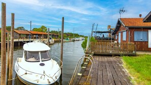A wooden dock with a boat moored to it, a wooden fence, and a building with a red roof in the background. - Apartment 'Chalet B Village Vacances' with Shared Pool, Private Terrace and Wi-Fi (Gujan-Mestras)