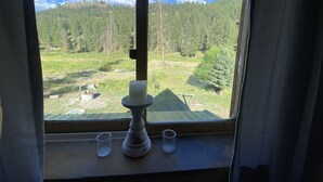 Ein Fenster mit Kerzenhalter und zwei Gläsern auf einem Holzboden, mit Blick auf eine bewaldete Landschaft und einen klaren Himmel.