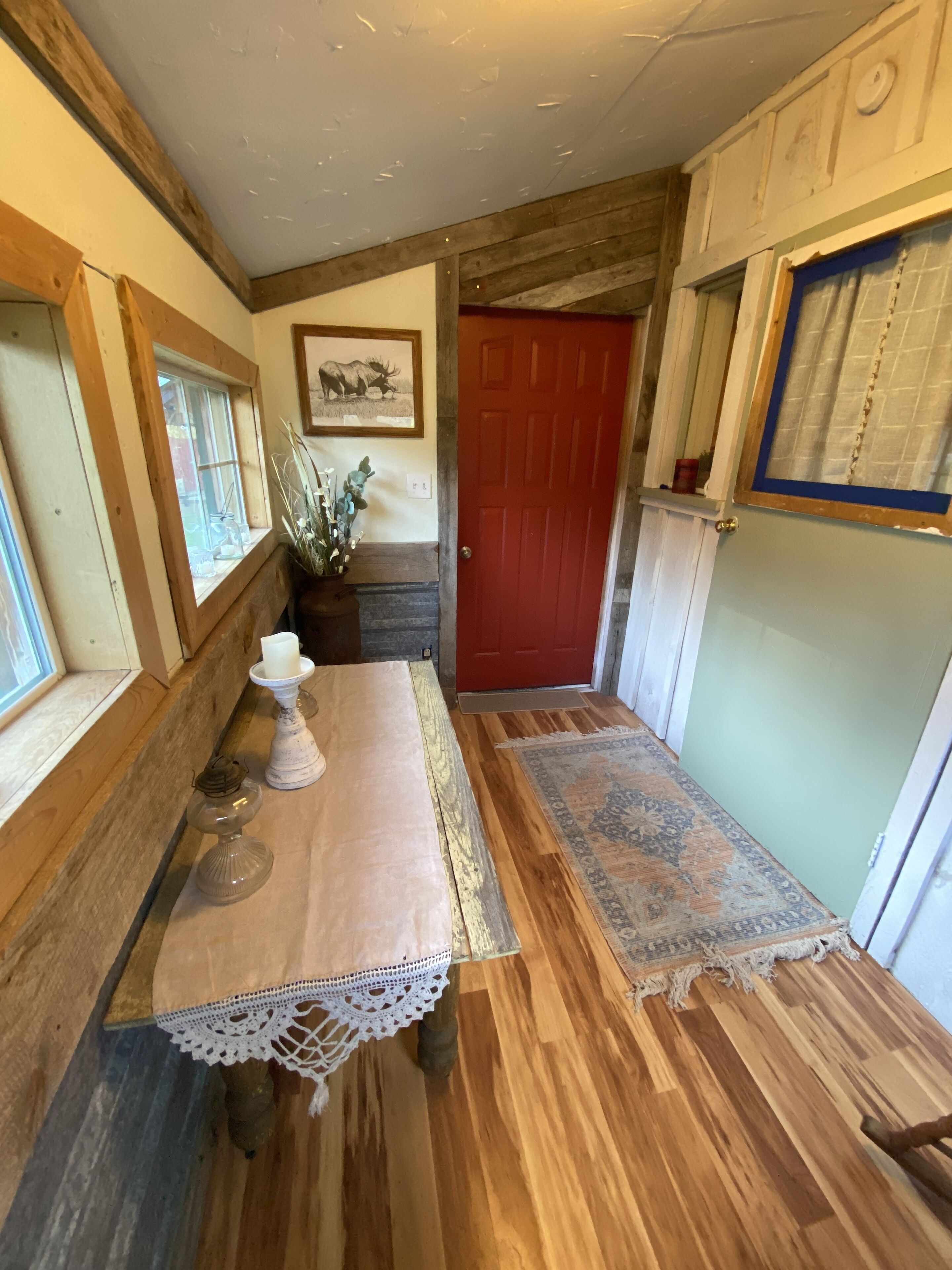 Un couloir étroit avec un plancher en bois, une porte rouge, une petite table recouverte d’une nappe en dentelle et un tableau encadré accroché au mur.