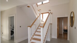 A spacious hallway with a wooden staircase leading to an upper floor. A washing machine is visible in the left corner, and a mirror hangs on the right wall.