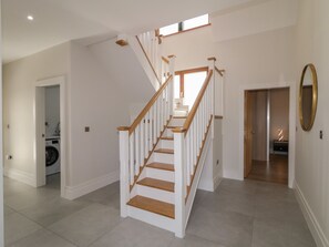 Ein geräumiger Flur mit einer Holztreppe, die zu einer oberen Etage führt. In der linken Ecke ist eine Waschmaschine zu sehen, und an der rechten Wand hängt ein Spiegel.