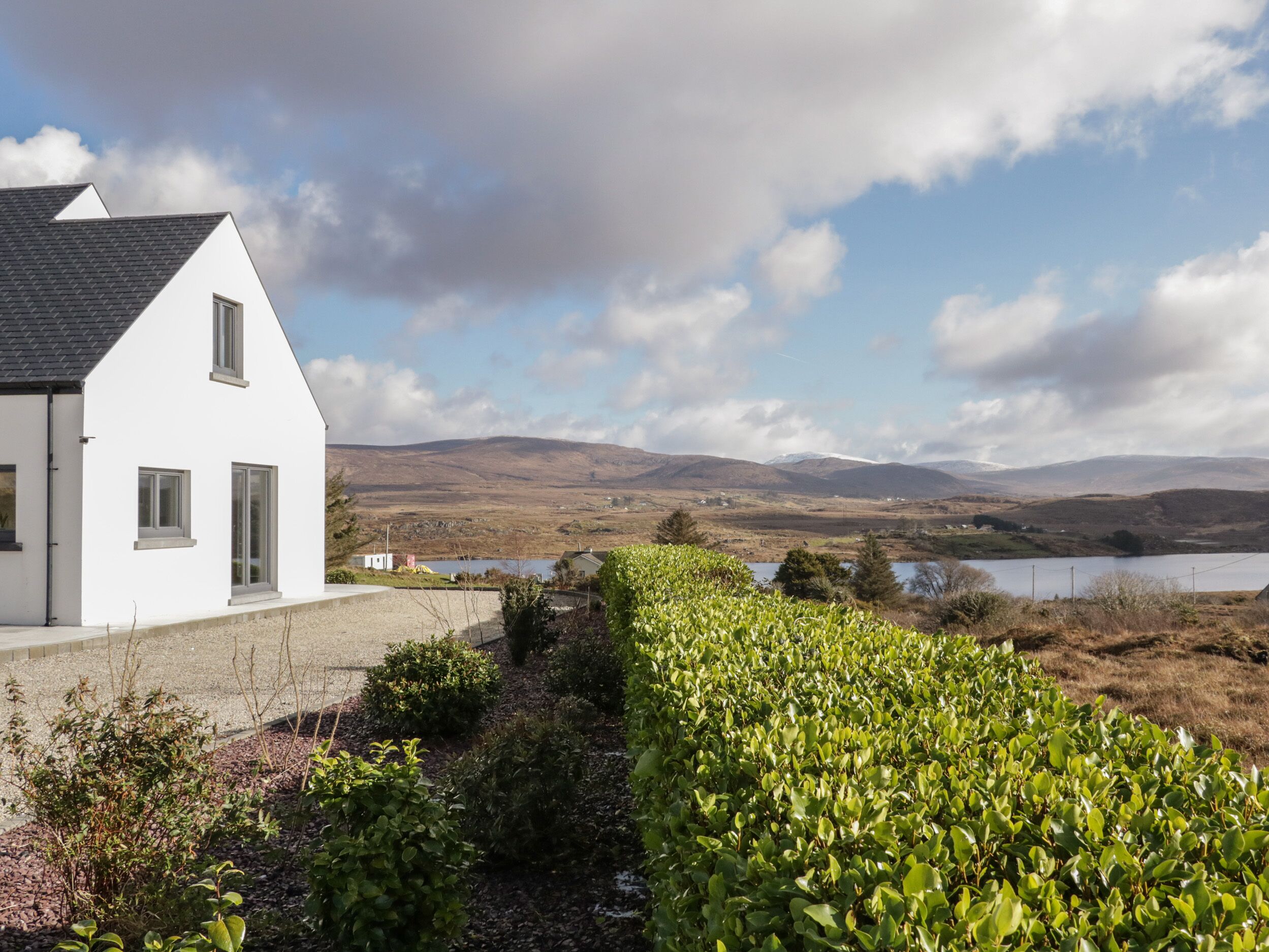 A white house with a sloped roof, a gravel driveway, and a well-maintained hedge.