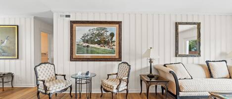 Una sala de estar con piso de madera, paredes blancas con paneles, un sofá, dos sillones, una mesa de centro y un cuadro.