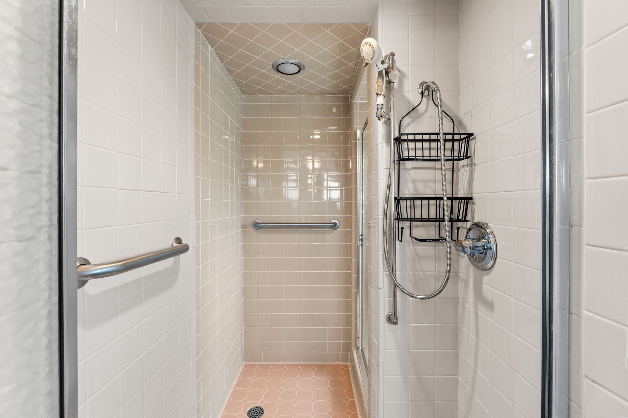 A small, enclosed shower area with white tiled walls, a glass door, a handrail, and a showerhead mounted on the wall.