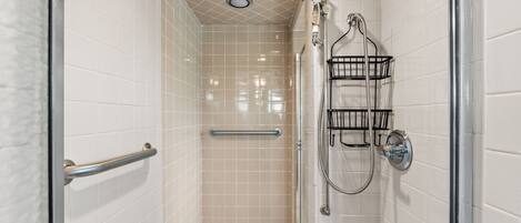 A small, enclosed shower area with white tiled walls, a glass door, a handrail, and a showerhead mounted on the wall.