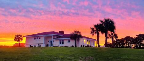A two-story house with a front porch, surrounded by a grassy lawn and palm trees.