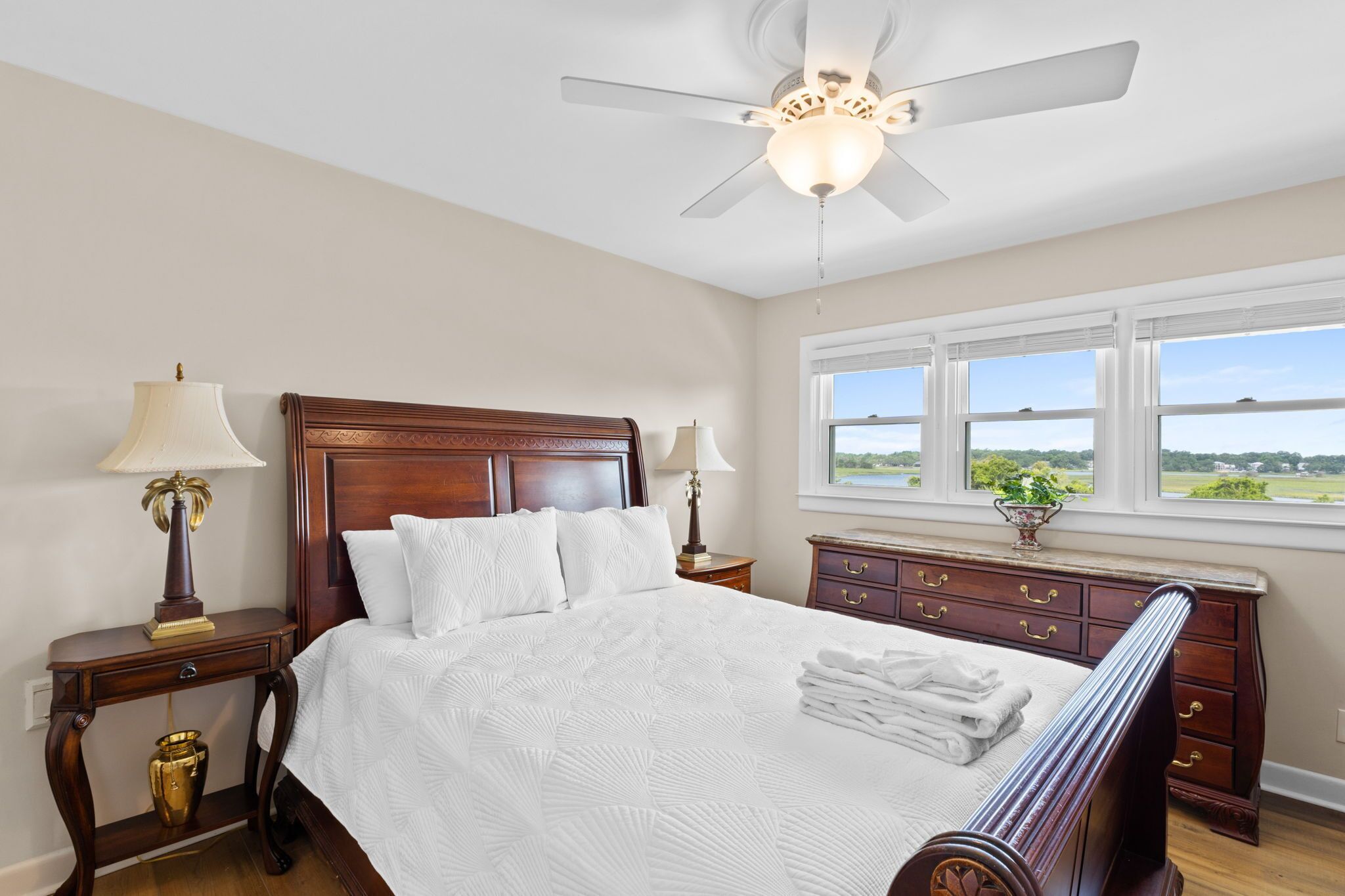 A bedroom with a large bed, two bedside tables, a nightstand, a dresser, a ceiling fan, and a window with a view of the outdoors.