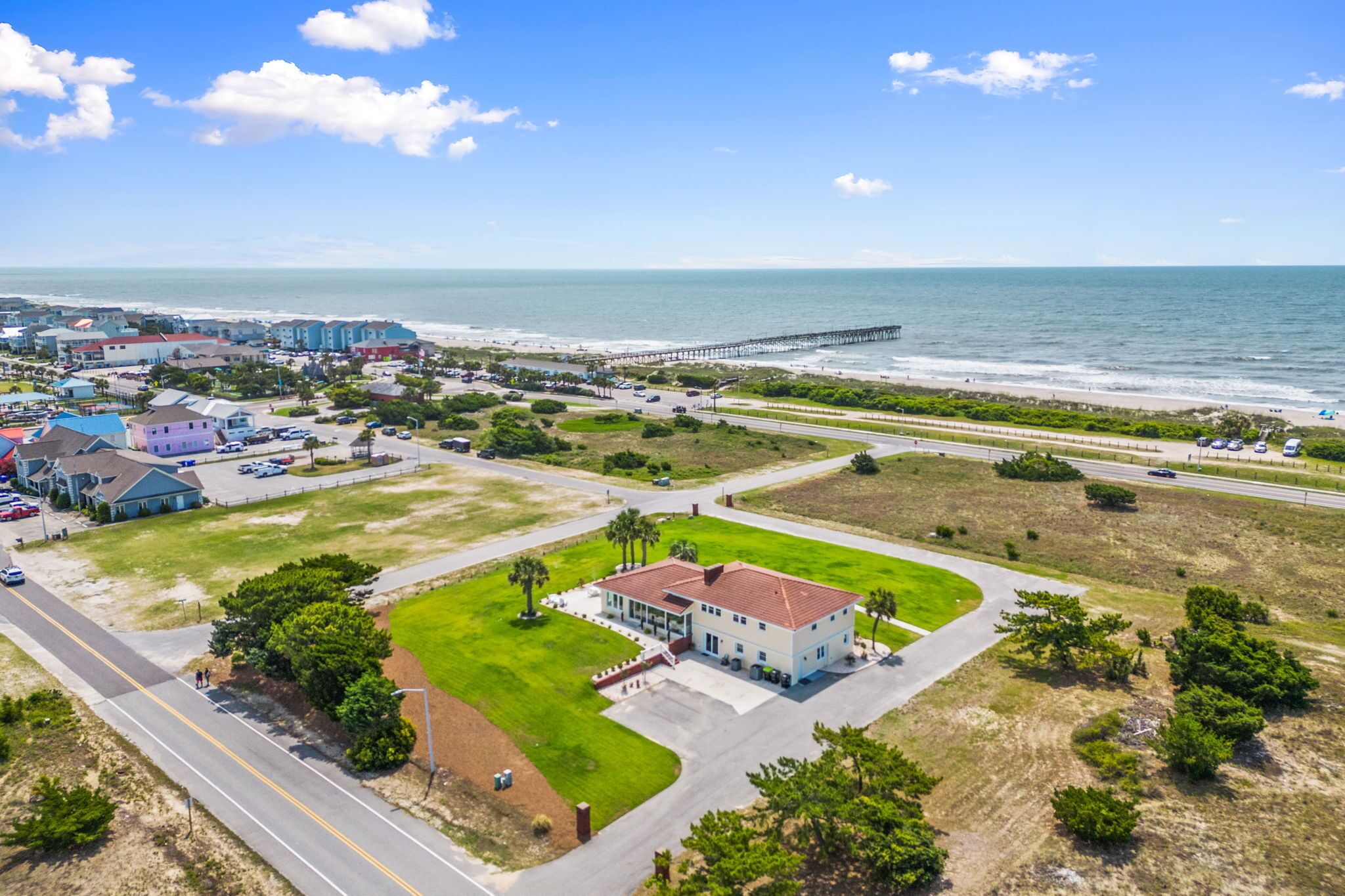 An aerial view of a coastal town with a prominent pier extending into the ocean, a large building with a red roof, and a well-maintained grassy area in the foreground.