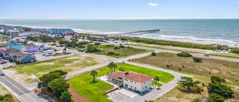 An aerial view of a coastal town with a prominent pier extending into the ocean, a large building with a red roof, and a well-maintained grassy area in the foreground.