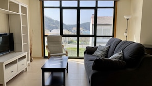 A living room with a sofa, a coffee table, a TV on a stand, a chair, and a large window with a view of buildings and hills. - Apartment with ocean views and community pool, just a 3 min walk to the beach! (O Porto de Espasante)