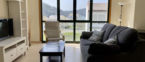 A living room with a sofa, a coffee table, a TV on a stand, a chair, and a large window with a view of buildings and hills.