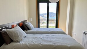 A bedroom with two beds, a window with a view of buildings and hills, and a radiator on the wall. - Apartment with ocean views and community pool, just a 3 min walk to the beach! (O Porto de Espasante)