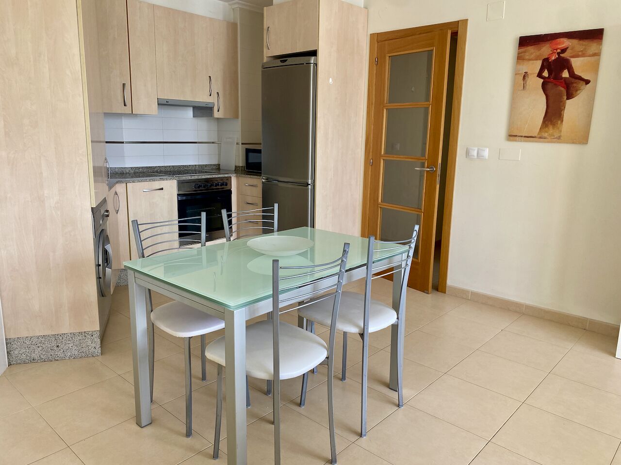 A modern kitchen with a glass dining table, stainless steel appliances, and light wooden cabinets.