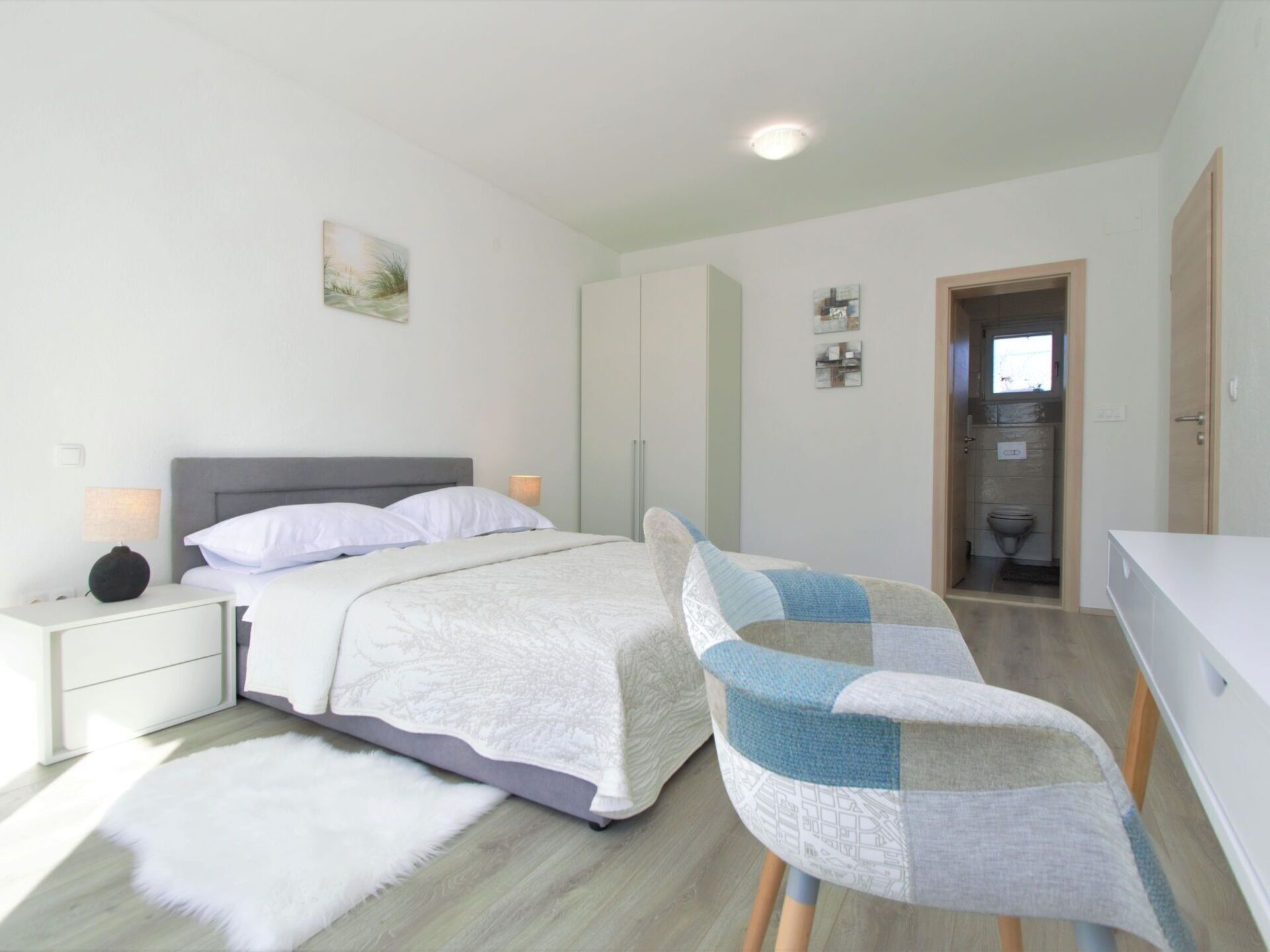 Een slaapkamer met een bed, een nachtkastje, een stoel, een wit tapijt en een badkamer zichtbaar door een geopende deur.