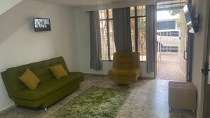 A living room with a green sofa, a yellow armchair, and a small ottoman. There is a green rug on the floor, and a flat-screen TV mounted on the wall. - Gold Stadium (Medellín)