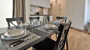 A dining area with a table set for four, including plates, glasses, cutlery, and napkins. There are four chairs around the table, and a kitchen is visible in the background.
