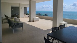 Un espace de détente extérieur moderne avec vue sur la mer, comprenant un coin salon avec une table et des chaises, ainsi qu’un espace de relaxation avec un fauteuil.