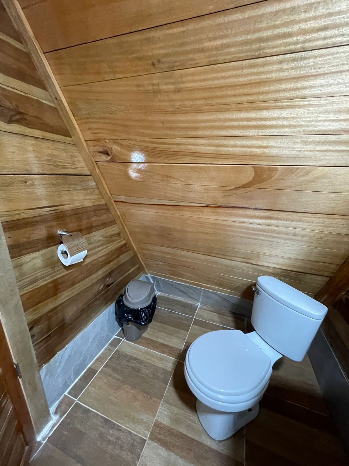 A wooden-paneled bathroom with a toilet, a small trash bin, and a toilet paper holder.