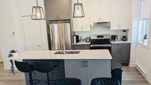 A modern kitchen with a central island, stainless steel appliances, and black bar stools. - Newly built condo, with excellent natural light, 20 minutes from the airport. (Sainte-Catherine)