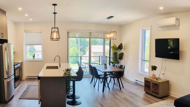 A modern kitchen with a central island, dining area with chairs, a sofa, and a TV mounted on the wall. - Newly built condo, with excellent natural light, 20 minutes from the airport. (Sainte-Catherine)