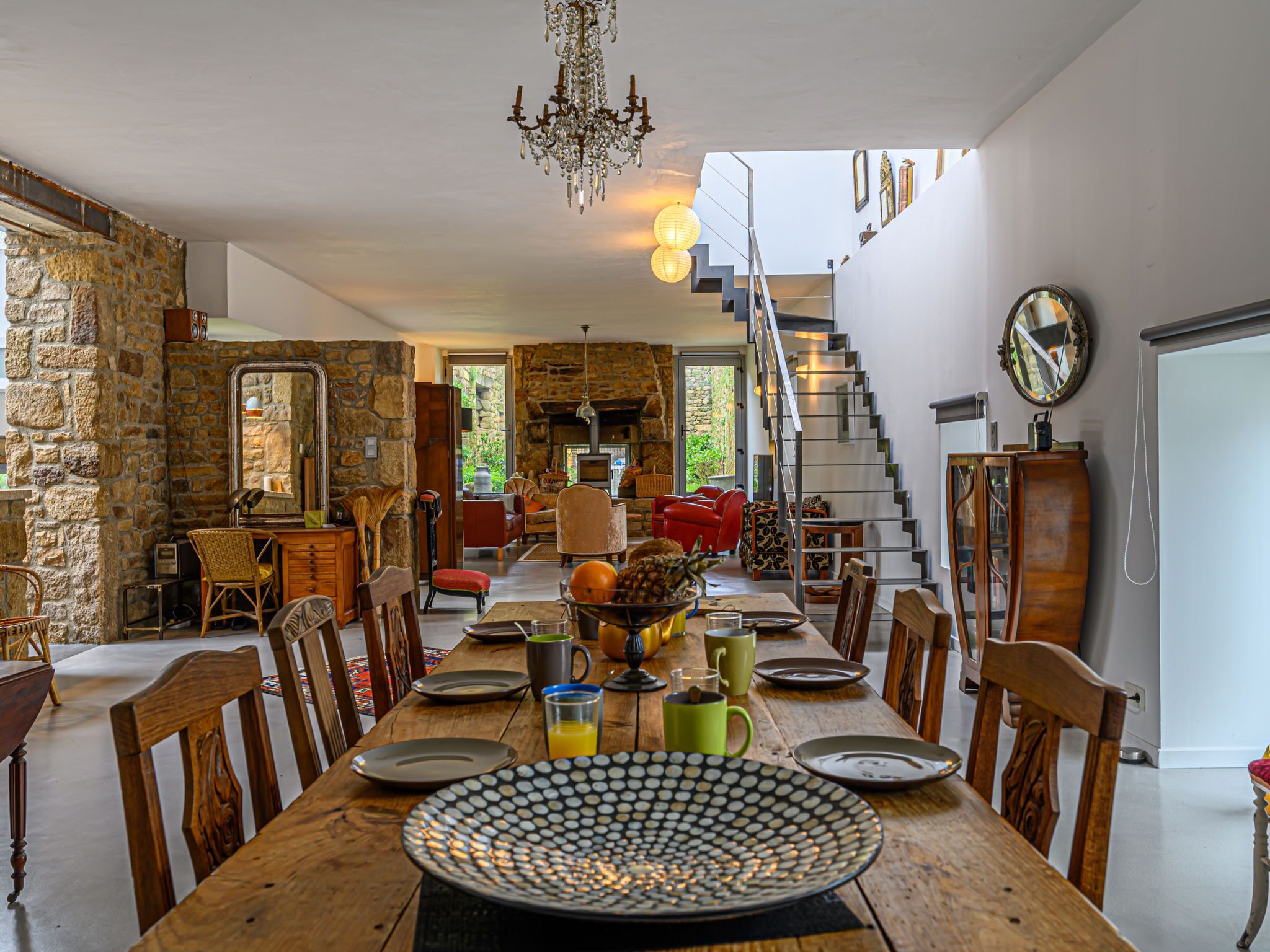 Un coin repas avec une table en bois, dressée pour quatre personnes, entourée de chaises. Un lustre est suspendu au plafond, et un escalier mène à un niveau supérieur.