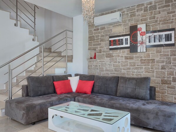 A modern living room with a grey sofa, red and white cushions, a glass coffee table, a staircase with metal railings, and a brick wall with two framed artworks. - Vacation home Bella by Interhome (Pula/Marčana)