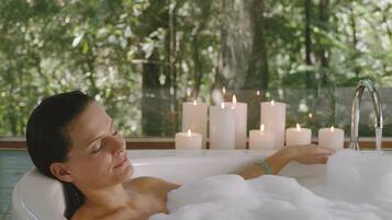 A person in a bathtub filled with bubbles, surrounded by lit candles, with a view of trees through a window.