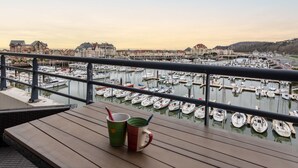 Une terrasse en bois avec deux tasses et une cuillère, donnant sur une marina où se trouvent de nombreux bateaux et, au loin, des bâtiments.