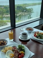 Una mesa de desayuno con diversos platos, un vaso de zumo de naranja y vistas a un lago a través de amplios ventanales.