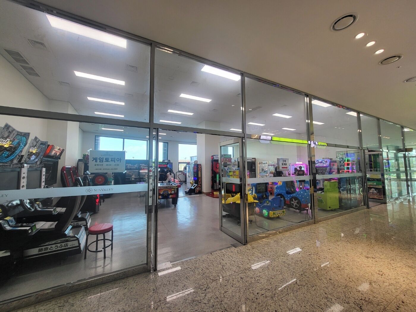 Eine moderne Indoor-Spielhalle mit Glaswänden, verschiedenen Spielautomaten und einer Sitzgelegenheit.