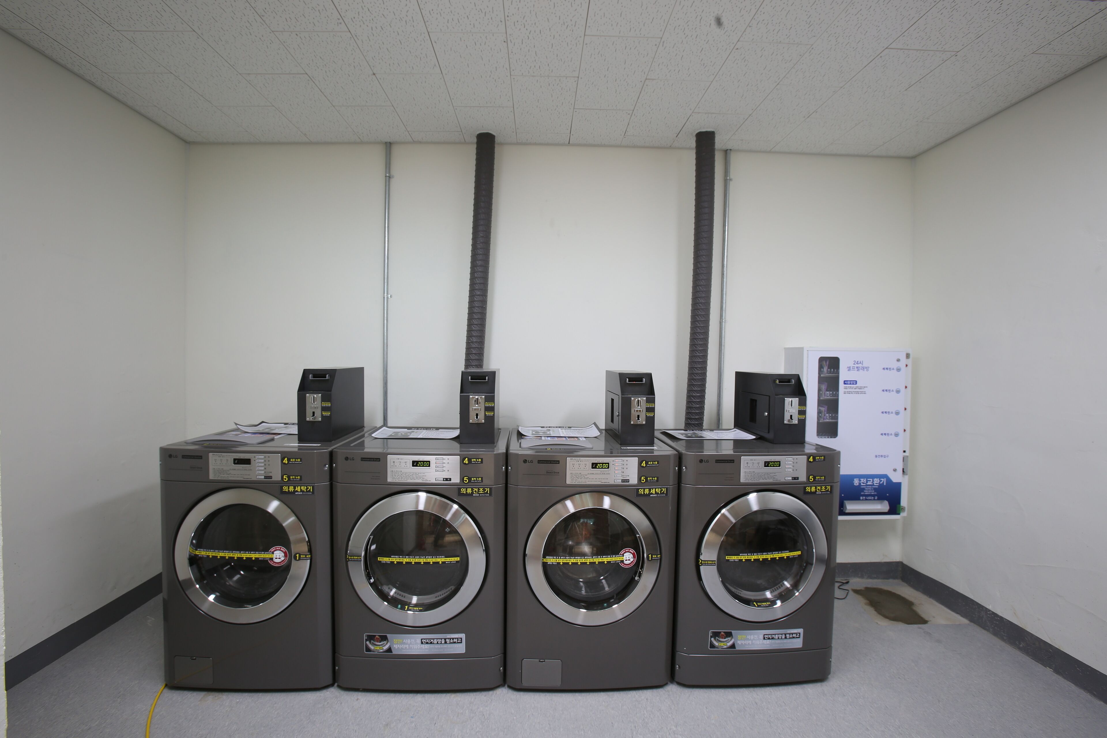 Four front-loading washing machines in a row with control panels on top, in a room with plain white walls and a tiled floor.