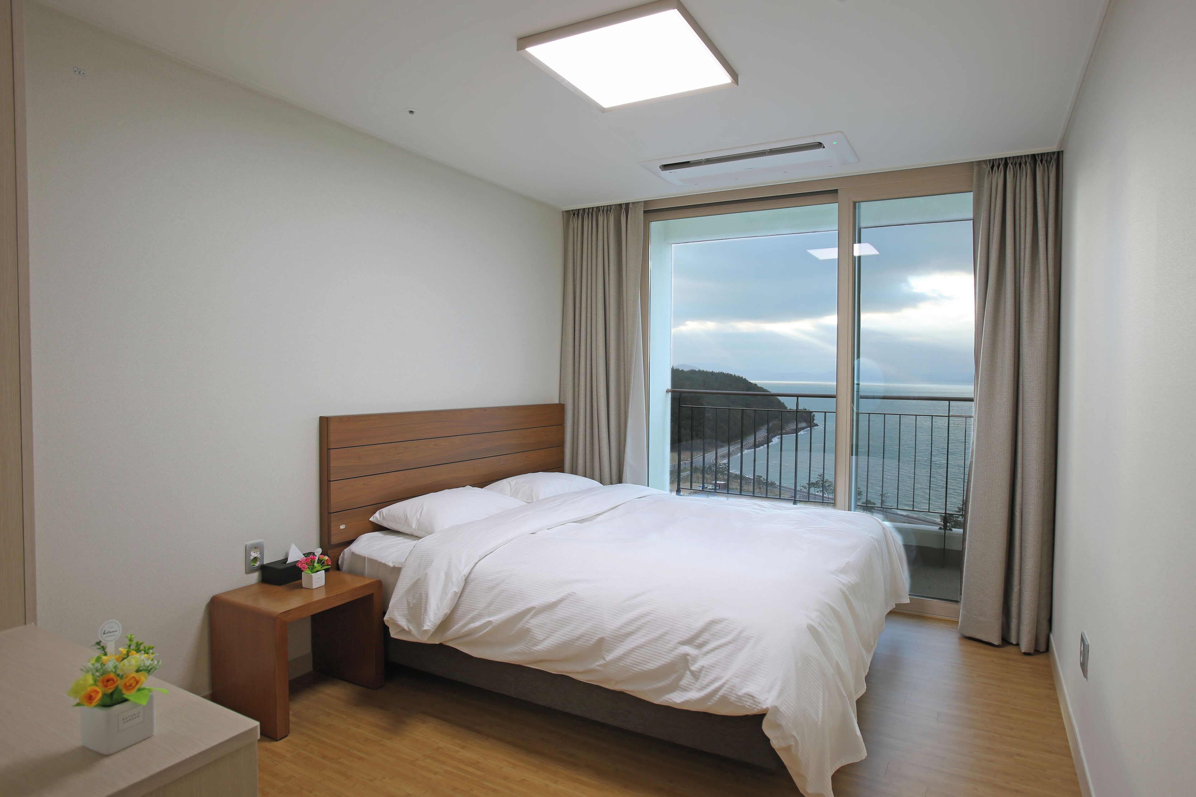 Habitación de hotel con cama, mesita de noche, una pequeña mesa con un florero y un gran ventanal con vistas al mar.