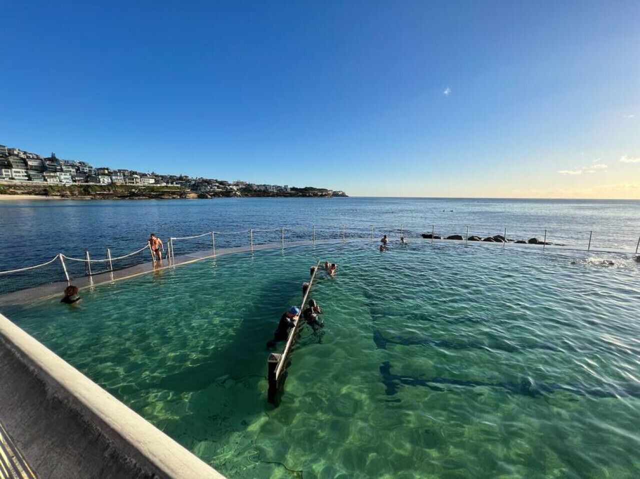 A clear pool with a wooden deck, surrounded by a rope barrier, and a coastal town in the background.