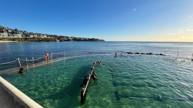 A clear pool with a wooden deck, surrounded by a rope barrier, and a coastal town in the background.