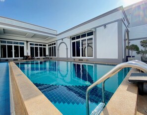 A rectangular indoor swimming pool with clear blue water, surrounded by a white building with large windows and a covered patio area. - Davinci Pool Villa (Pattaya)