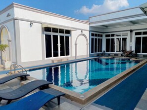 A swimming pool with lounge chairs, a covered area with a table, and a building with large windows and columns. - Davinci Pool Villa (Pattaya)