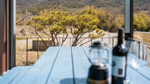 Una mesa de madera con una botella y un vaso, con vista a un árbol y un paisaje montañoso.