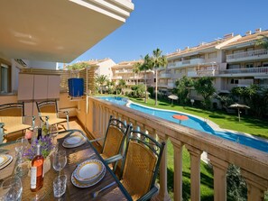 A balcony with a dining table set for two, overlooking a swimming pool and a well-maintained garden.