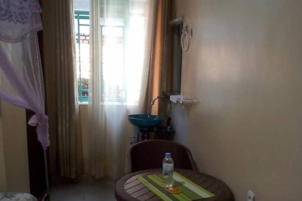 Una habitación pequeña y tenuemente iluminada, con una cama, una mesa y una botella de agua, además de un ventanal con cortinas.