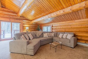 A log cabin interior with a large L-shaped sectional sofa, a small coffee table, and a lamp on a side table.