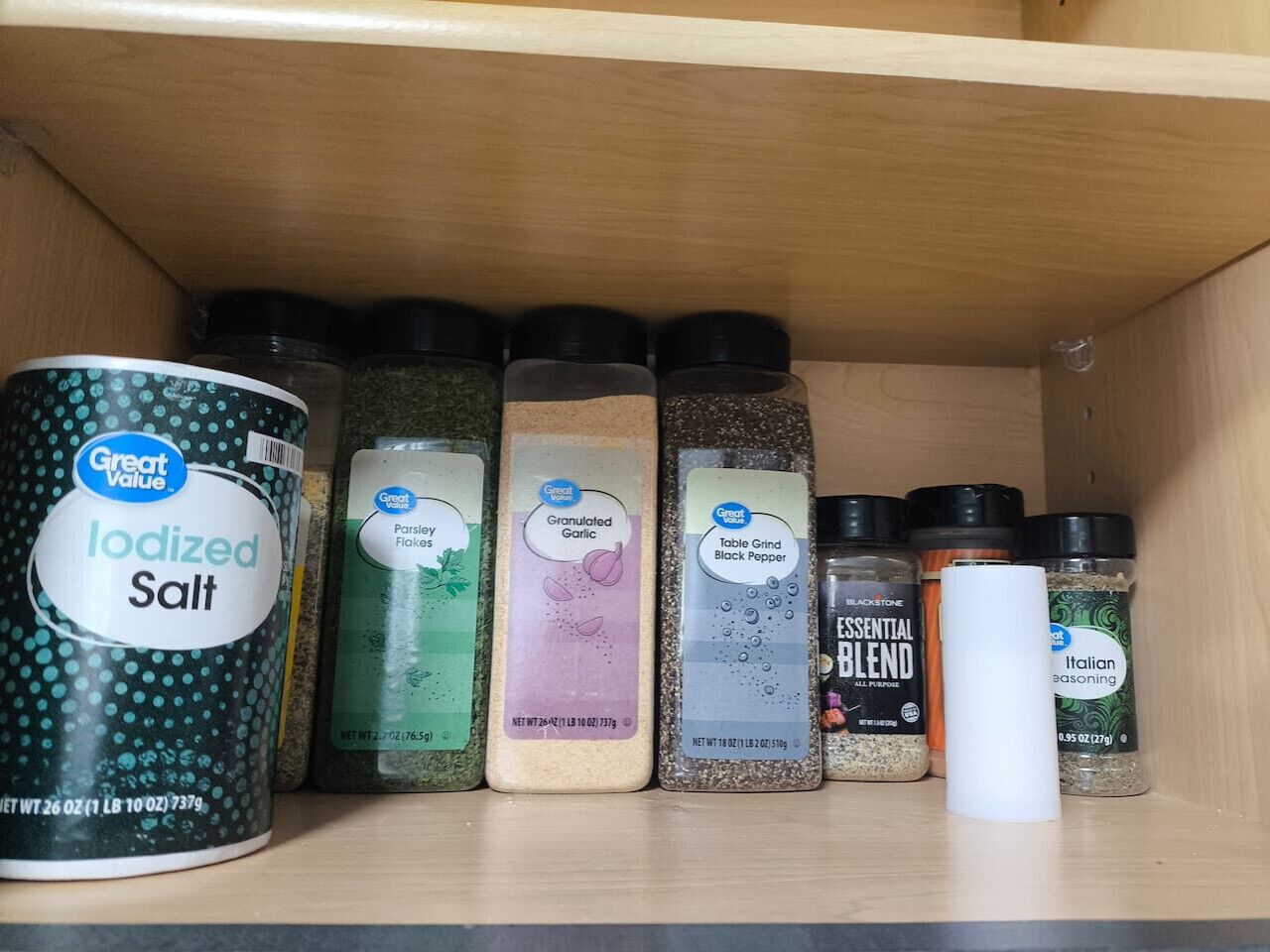 A shelf with various spice containers including iodized salt, parsley flakes, granulated garlic, black pepper, and Italian seasoning.