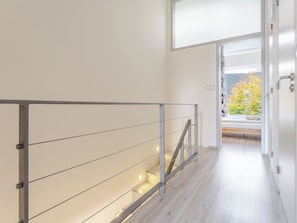 Un intérieur moderne avec un plancher en bois, une rambarde métallique dotée de lumières et une vue dégagée sur l’extérieur grâce à une porte ouverte.