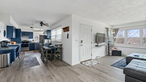 Eine moderne Küche mit blauen Schränken, einem Essbereich mit Tisch und Stühlen, einem Deckenventilator und einem großen Fenster mit Vorhängen.
