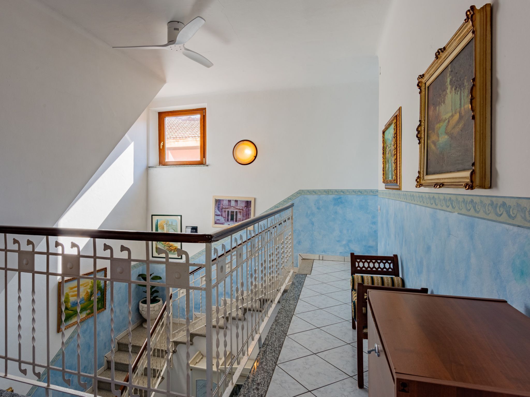 Una escalera con barandilla, un ventilador de techo, y una pared con cuadros y una pintura.