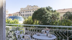 Un balcon avec des chaises et une table en métal peintes en blanc, une machine à café et des tasses. Au fond, on aperçoit un bâtiment sur lequel est apposée une enseigne indiquant « GRAND HÔTEL ALBERT ».