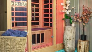 Eine Holzsauna mit Glastüren, ein Korbsessel mit blauem Handtuch und zwei Zimmerpflanzen-Orchideen.