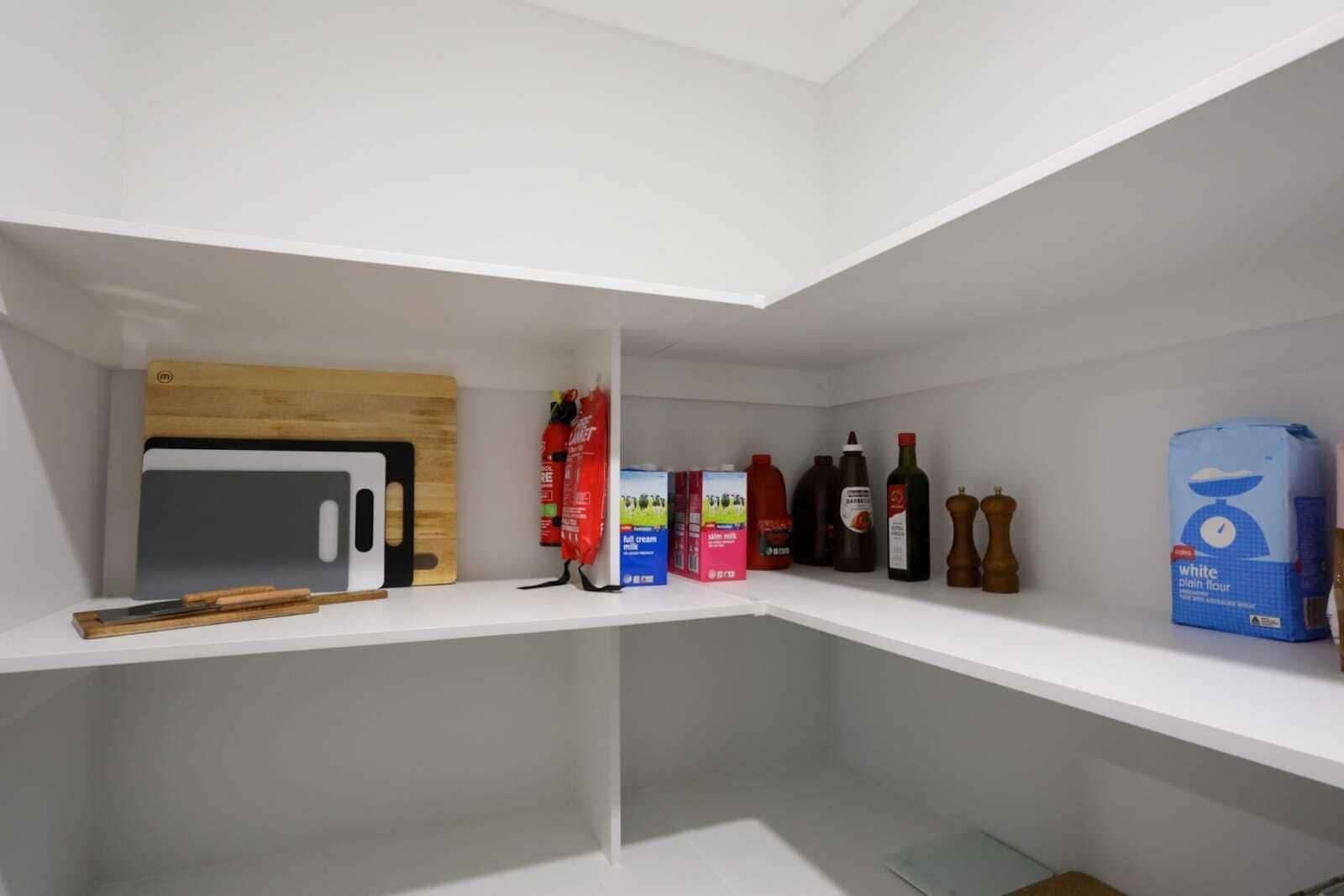 A kitchen with white shelves, a wooden cutting board, a knife, a carton of milk, a bottle of sauce, a bottle of olive oil, a bottle of vinegar, a bottle of wine, a bottle of cooking oil, a bag of flour, and two pepper mills.