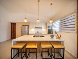 A modern kitchen with a central island, stainless steel appliances, wooden stools, and pendant lighting.