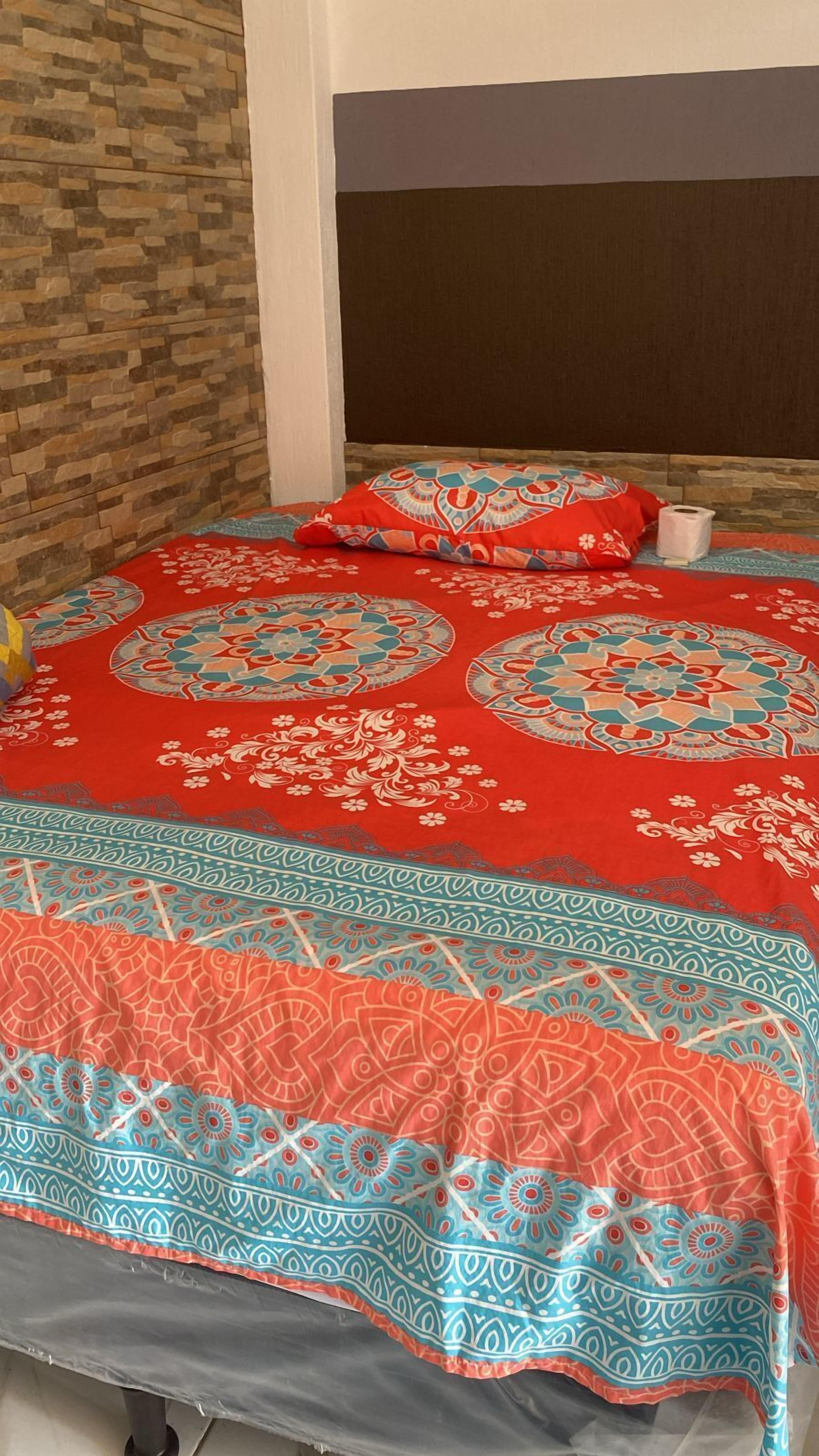 Una cama con una colcha estampada y almohadas, un rollo de papel higiénico y una pared con diseño de ladrillos.
