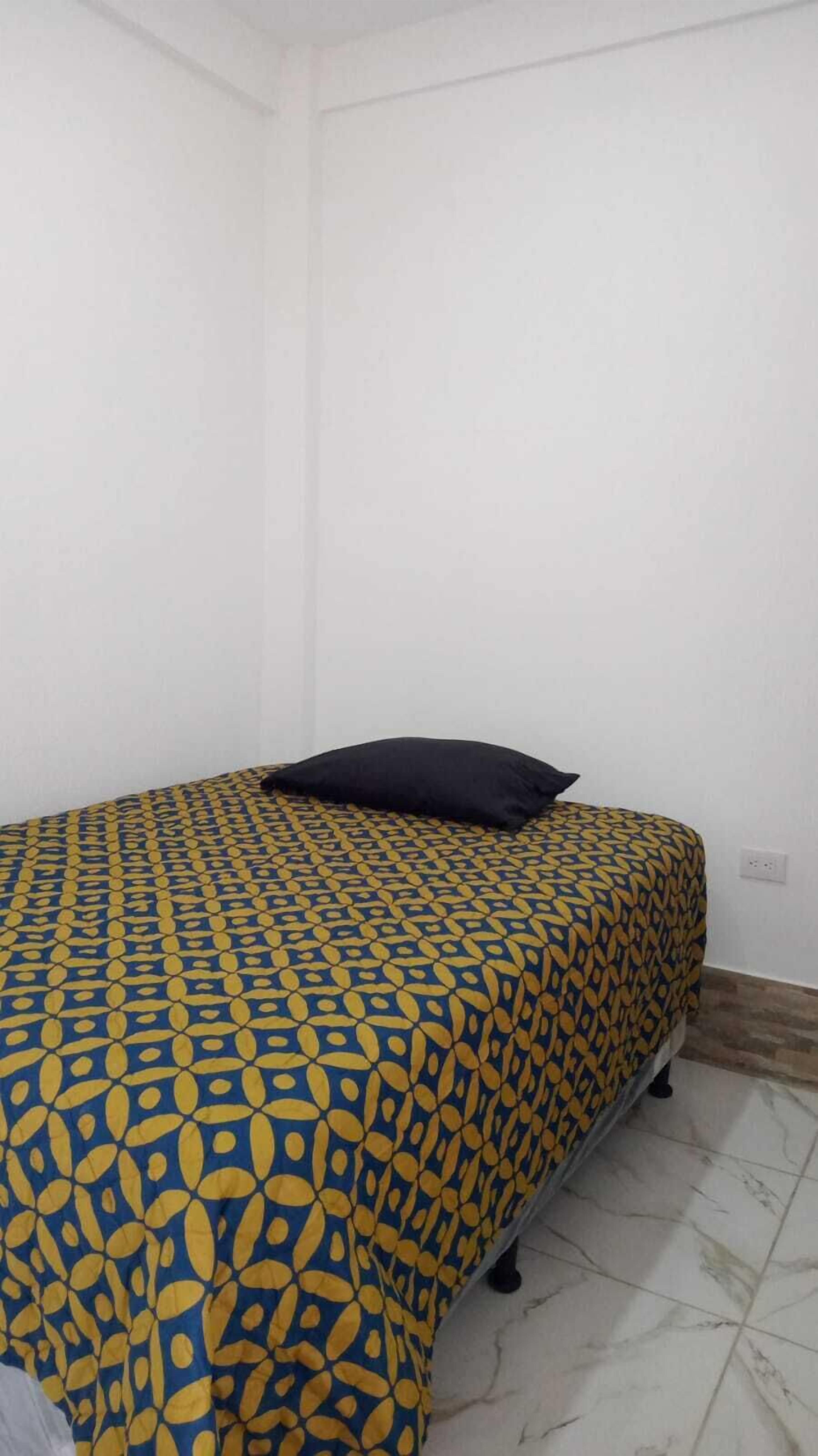 Ein Einzelbett mit einem gemusterten Bettüberwurf und einem schwarzen Kopfkissen in einem Zimmer mit weißen Wänden und Fliesenboden.